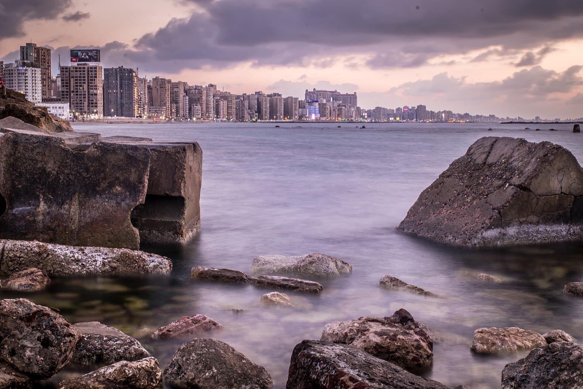 صخور الشاطئ عند بئر مسعود والبحر الهادئ، وعمارات الكورنيش على امتداد الأفق عند الغروب